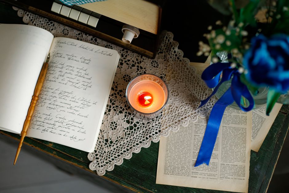 A cozy desk setting with a lit candle, open journal, lace doily, and blue floral bouquet.